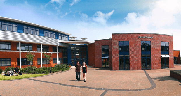 Two people are seen walking toward the viewer in front of a red brick building with the words ‘Bexhill College’ on the building. 