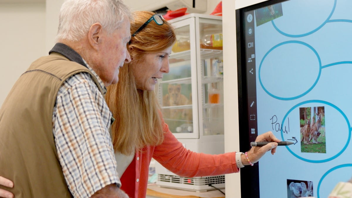 A resident at a senior home in Colorado, along with his aid, interacting with a brainstorming activity on the SMART Board.