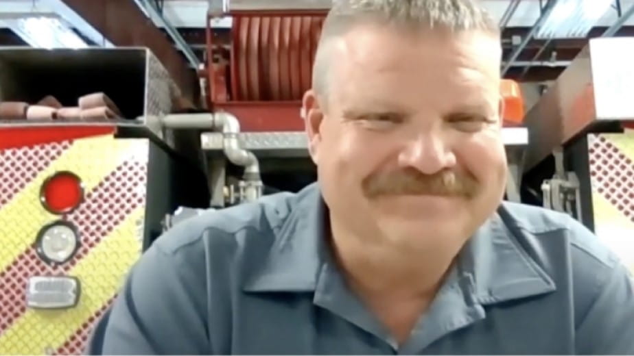 A smiling firefighter, sitting in a fire station, participates in a video call, showing his readiness and friendliness.