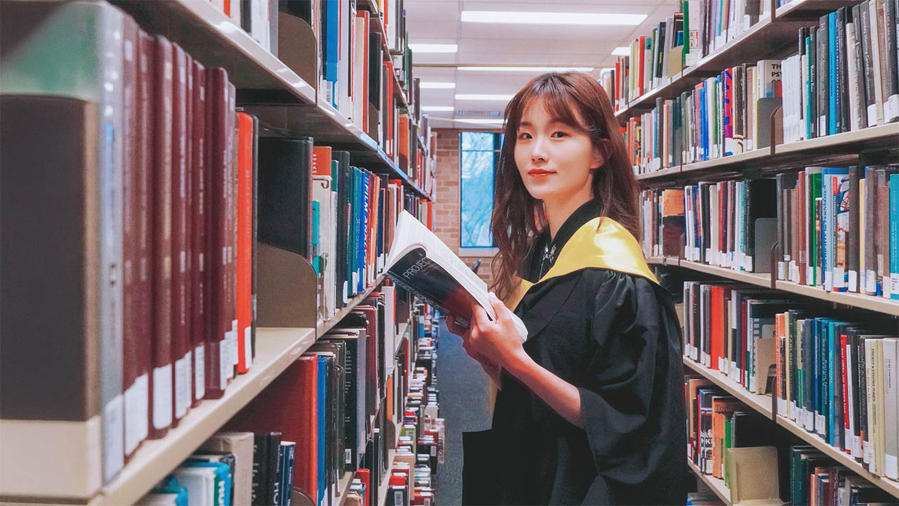 A graduate student holding a book while standing between rows of bookshelves in a library.