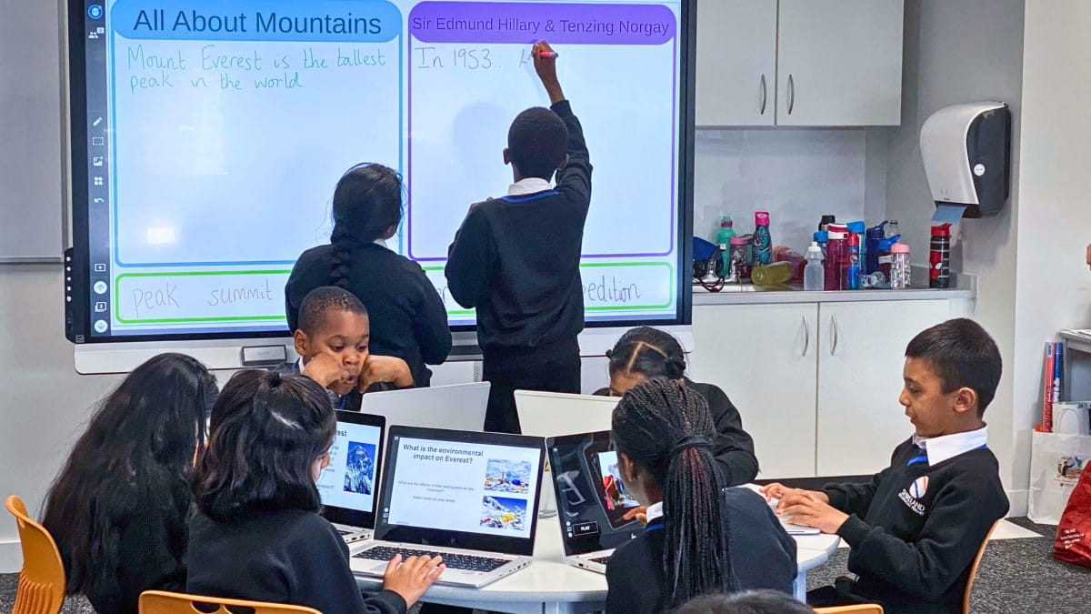 Two students working at a SMART interactive display, while the rest of their group works independently on their student devices at a table in front. 