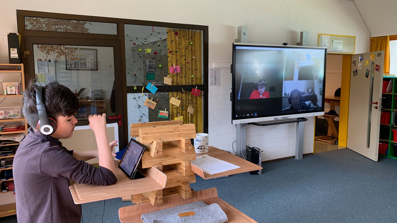 Student using a tablet with headphones to participate in an online class, with a SMART Board displaying classmates on a video call.