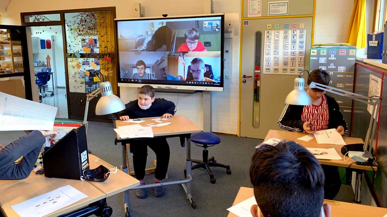 Hybrid classroom with students working at desks and engaging with remote peers displayed on a SMART Board.