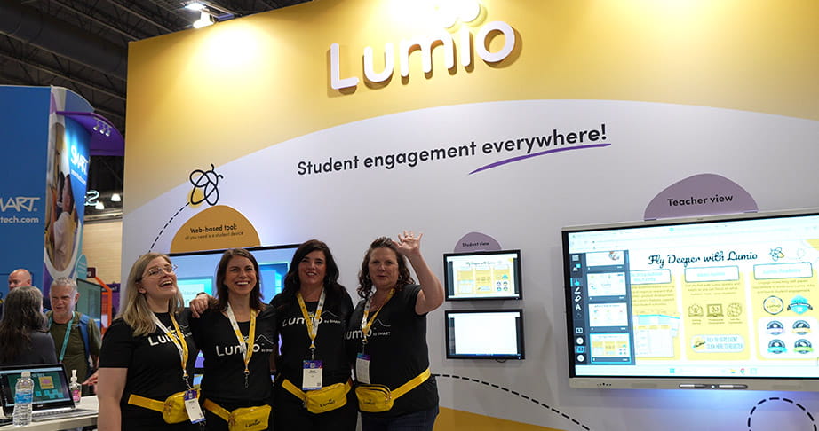 Four women, wearing Lumio t-shirts, smiling and standing in front of a Lumio booth that showcases educational tools for student engagement.