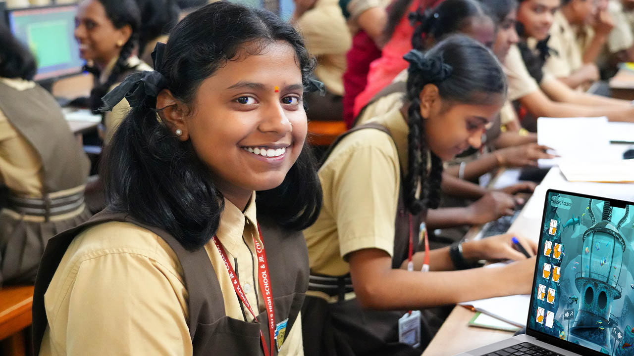 Student smiling while engaging in a laptop-based activity in a classroom, with classmates in the background.
