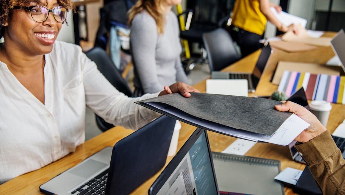 A woman in an office setting passing a document, with laptops and work materials around, indicating partnership.