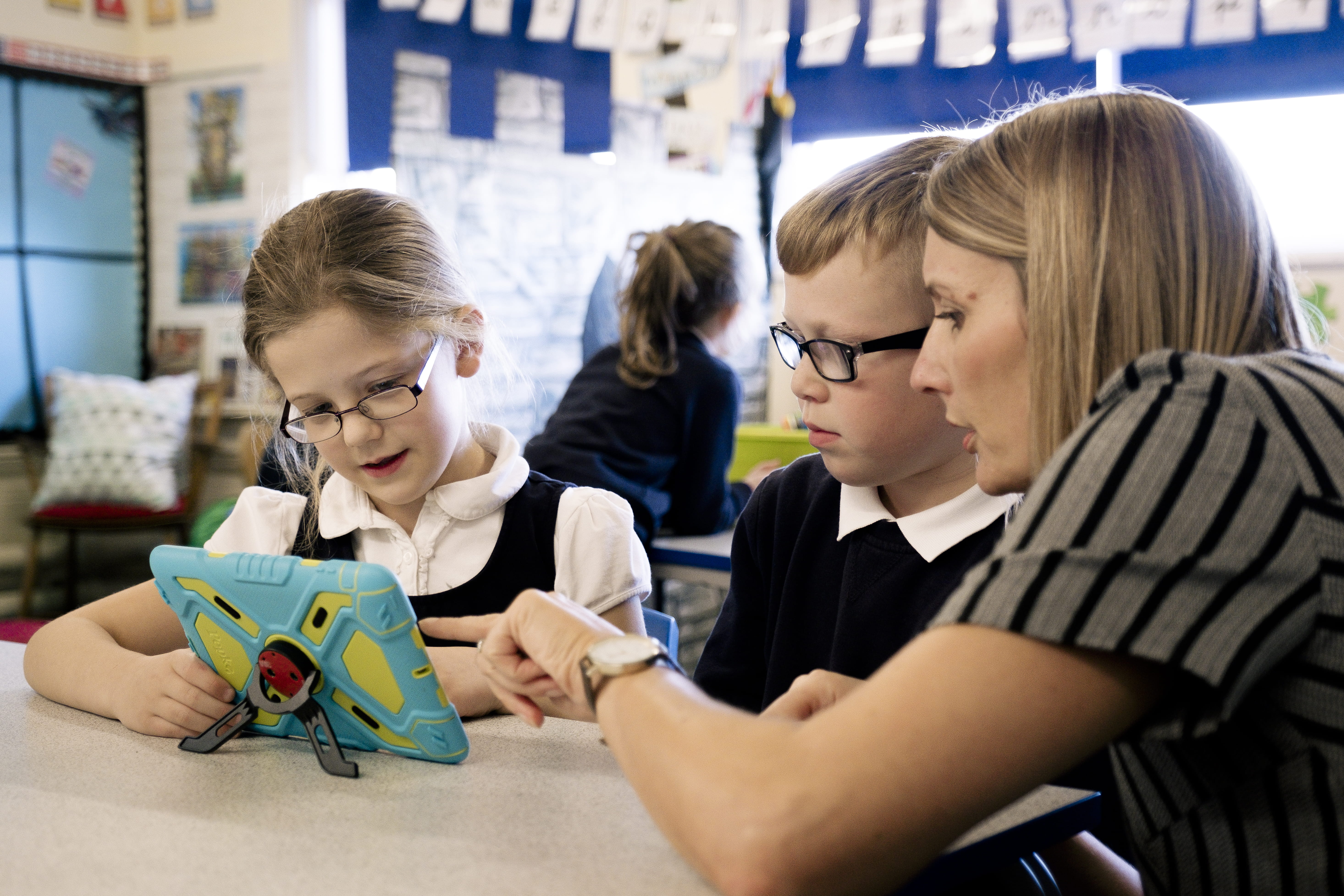 A teacher helps two students on a tablet
