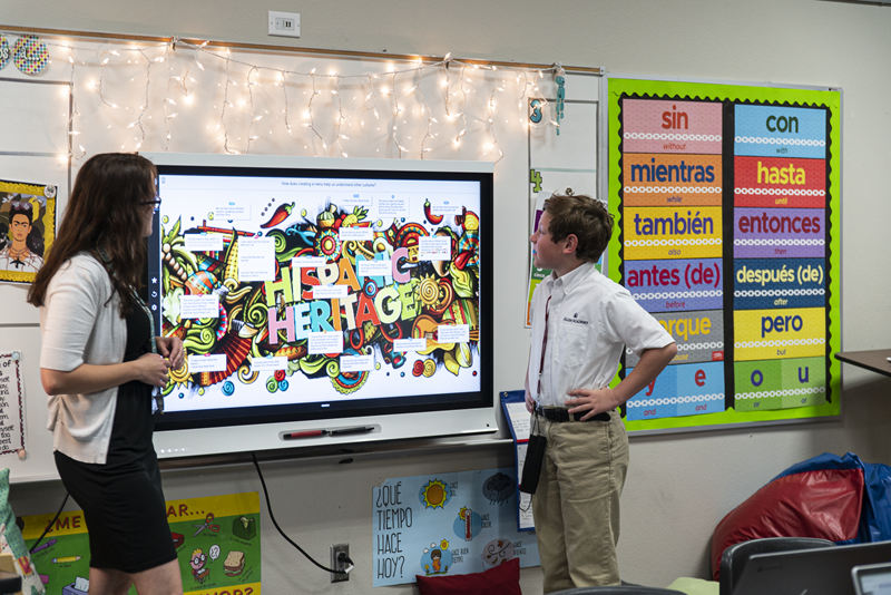 Teacher and student engaging with a SMART Board displaying Hispanic Heritage content in a decorated classroom.
