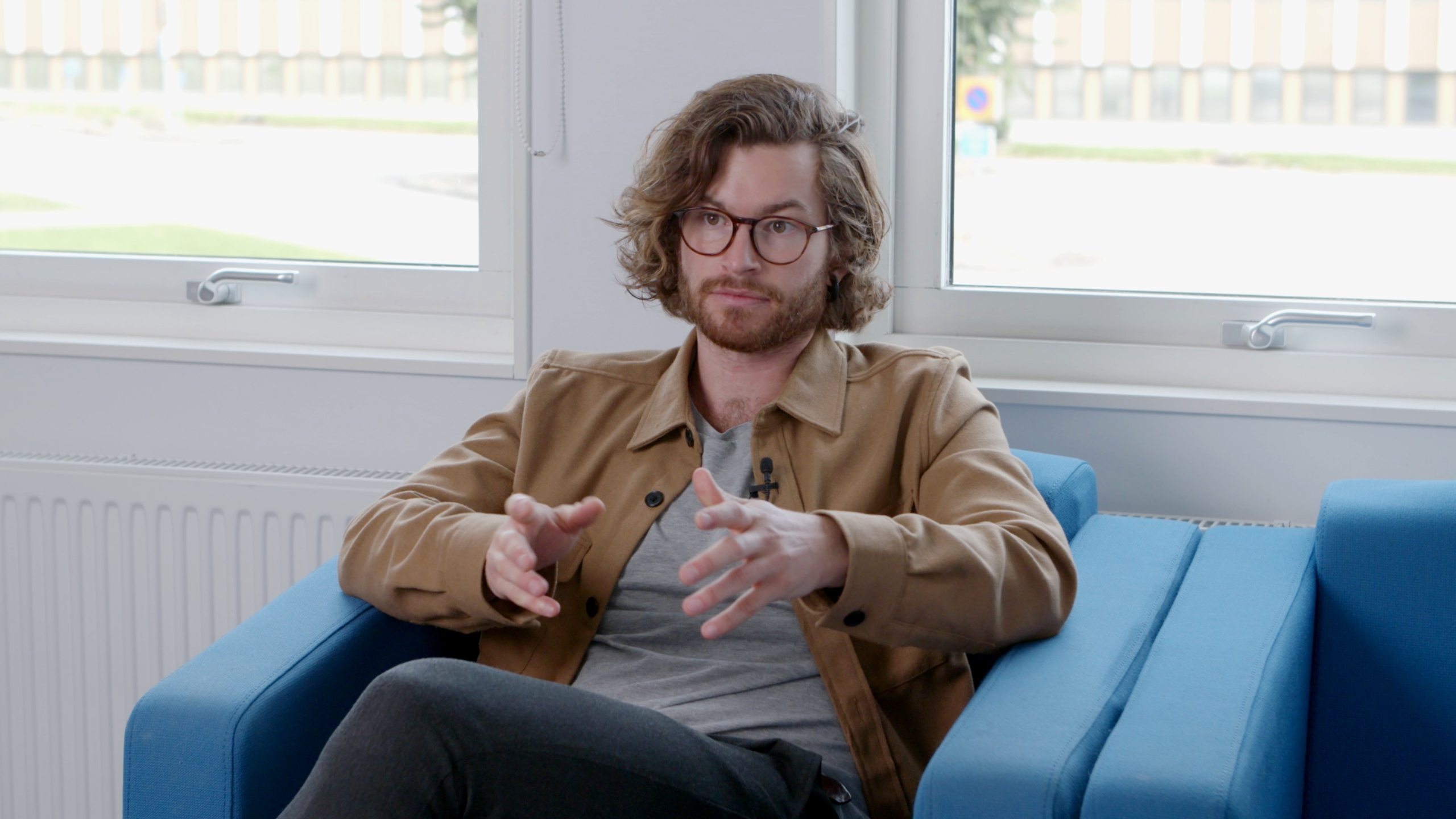 A male teacher sitting on a blue chair, explaining concepts during a video interview.