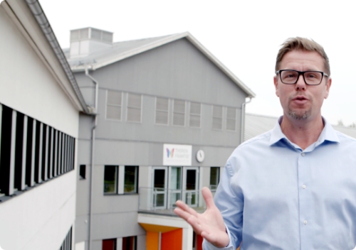 Educator explaining in front of a gray school structure with large windows.