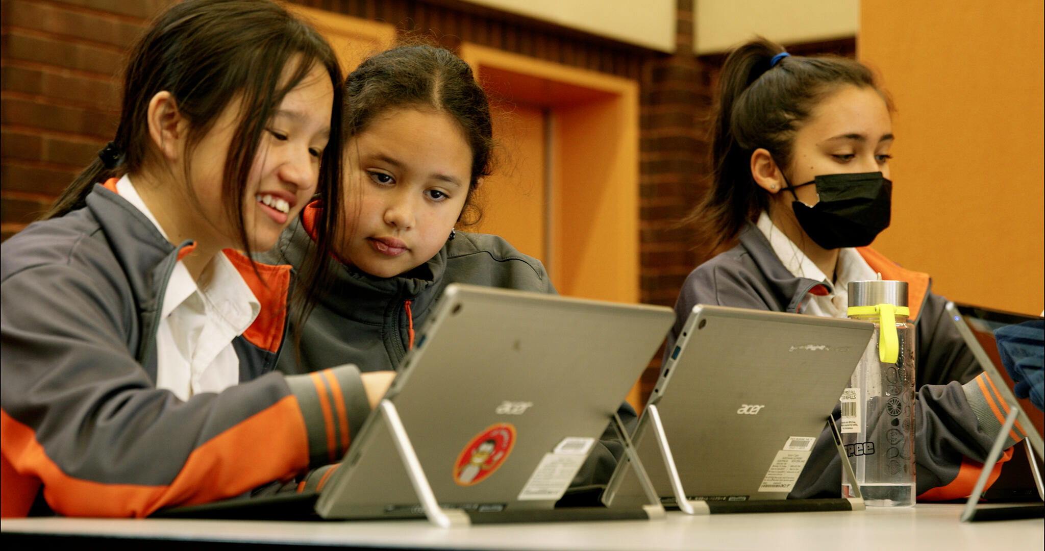 Three students collaborate on laptops during a classroom activity, with one wearing a mask.