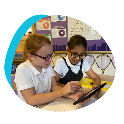 Two young students in white uniforms engrossed in collaborative learning on a tablet, seated in a classroom with educational posters in the background.